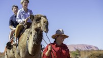 image of two people on camel being led by a guide with Uluru in background 